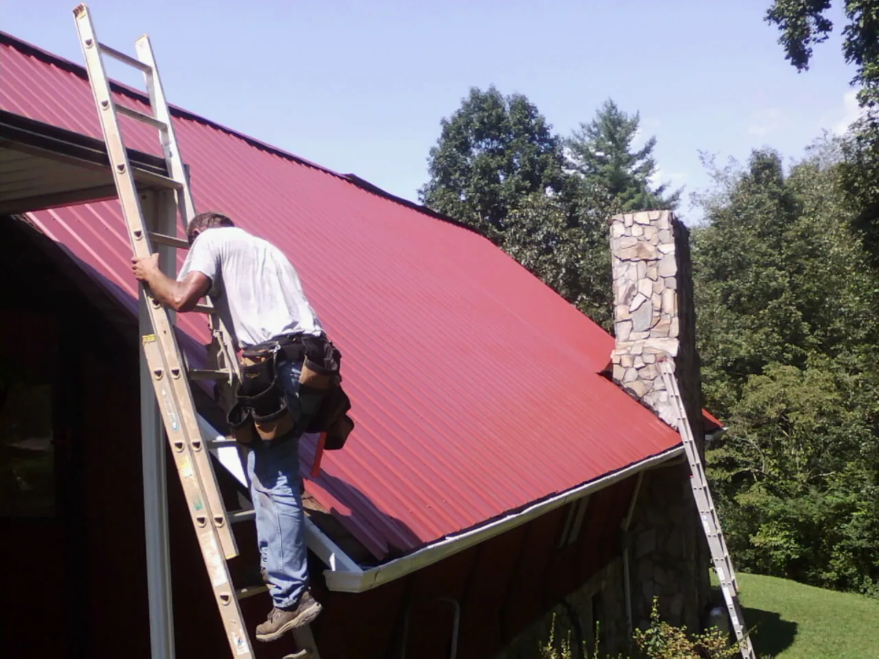 Roofer on a ladder beside a red metal roof and stone chimney — Soffit & Fascia Repair by Roofing Brothers in Braddock Heights